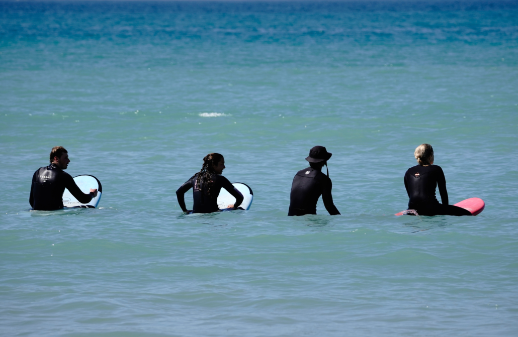 Cuatro personas con trajes de neopreno esperan en el agua con sus tablas de surf, mirando hacia el mar.