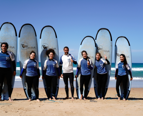 Grupo de siete personas con tablas de surf posando en la playa, con el mar al fondo y cielo azul.