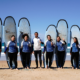 Grupo de siete personas con tablas de surf posando en la playa, con el mar al fondo y cielo azul.