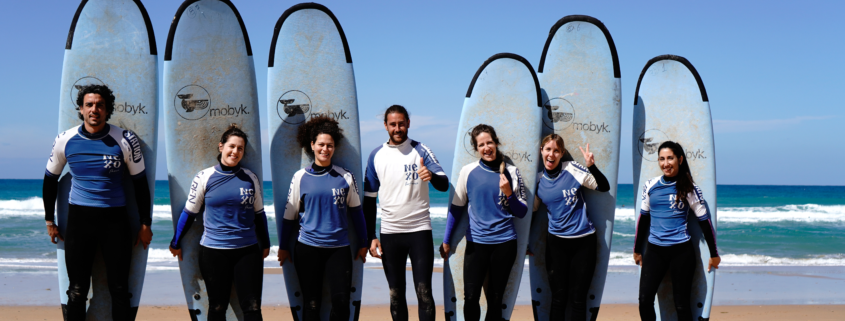 Grupo de siete personas con tablas de surf posando en la playa, con el mar al fondo y cielo azul.