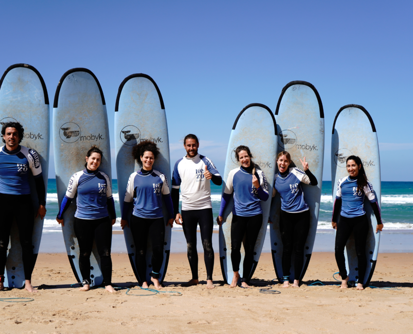 Grupo de siete personas con tablas de surf posando en la playa, con el mar al fondo y cielo azul.