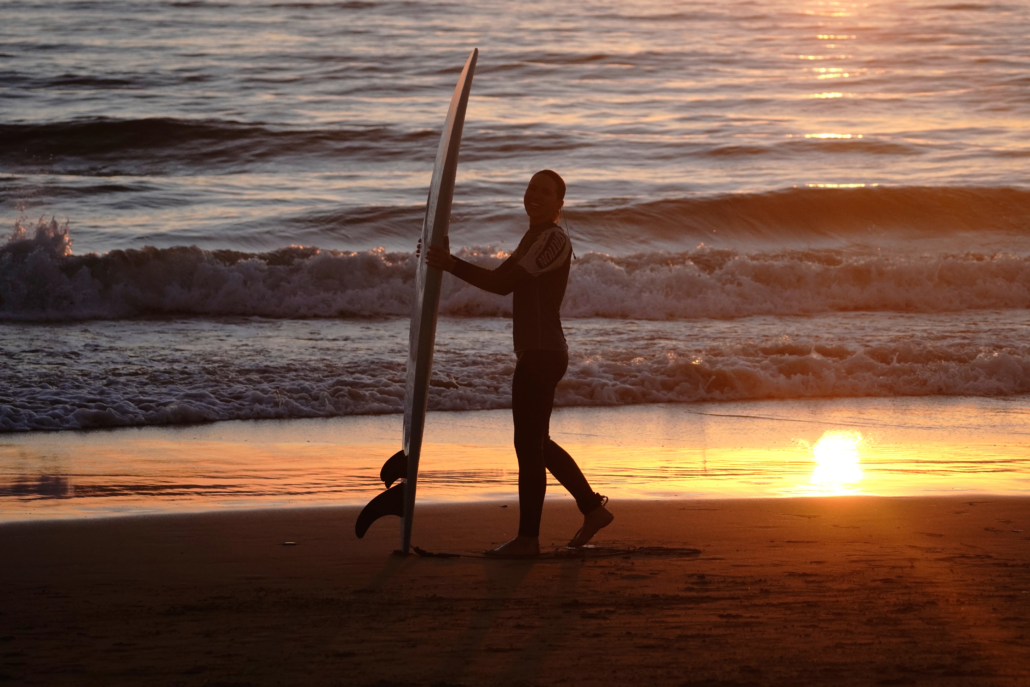 Surfista caminando por la orilla con su tabla al atardecer, con el mar y las olas de fondo.
